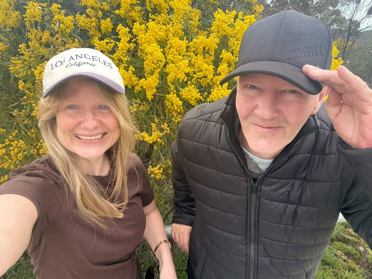 A support worker and participant smiling together beside flowering wattle on a sunny walk