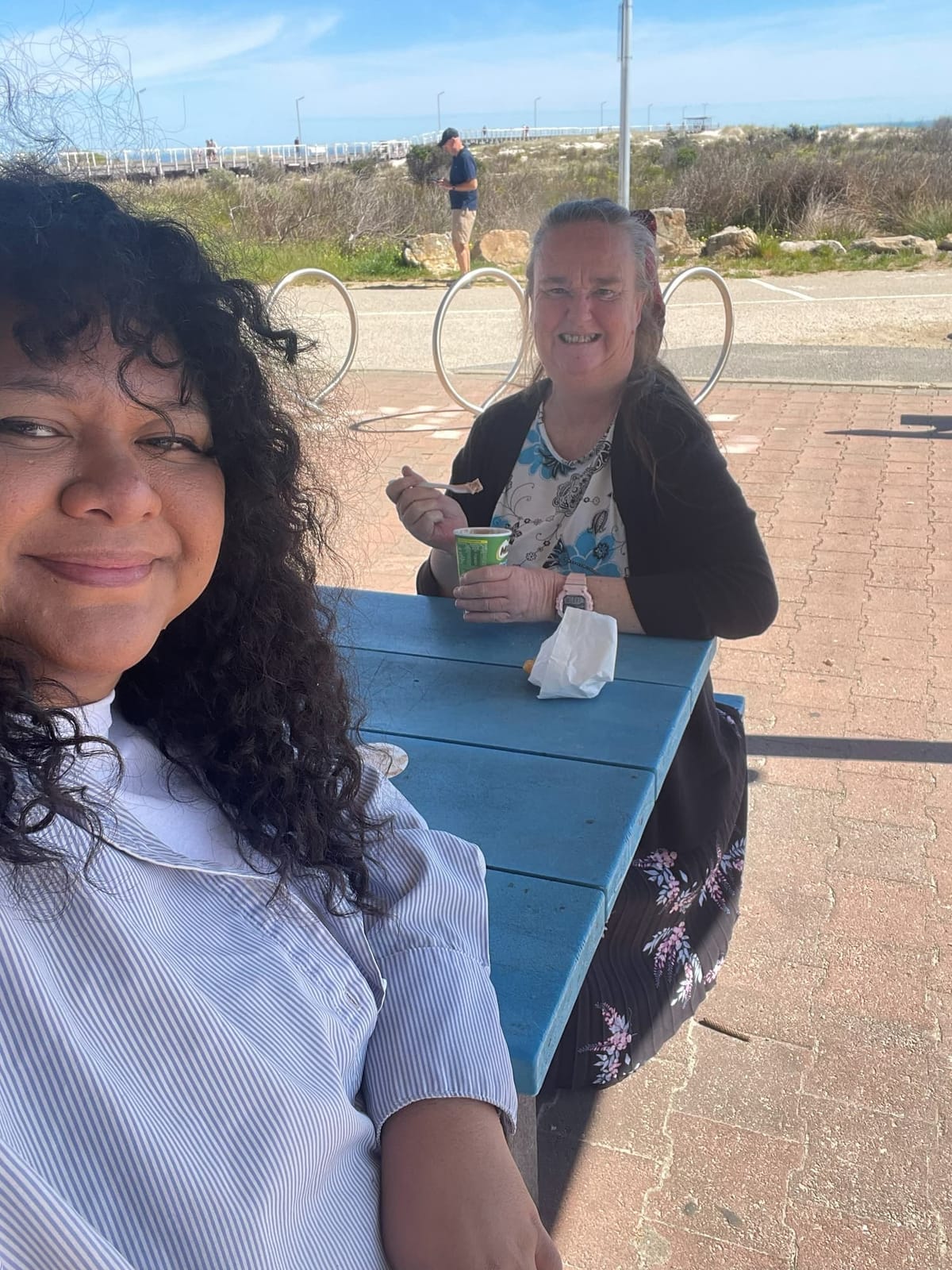 Two women smiling together at a blue table by the sea
